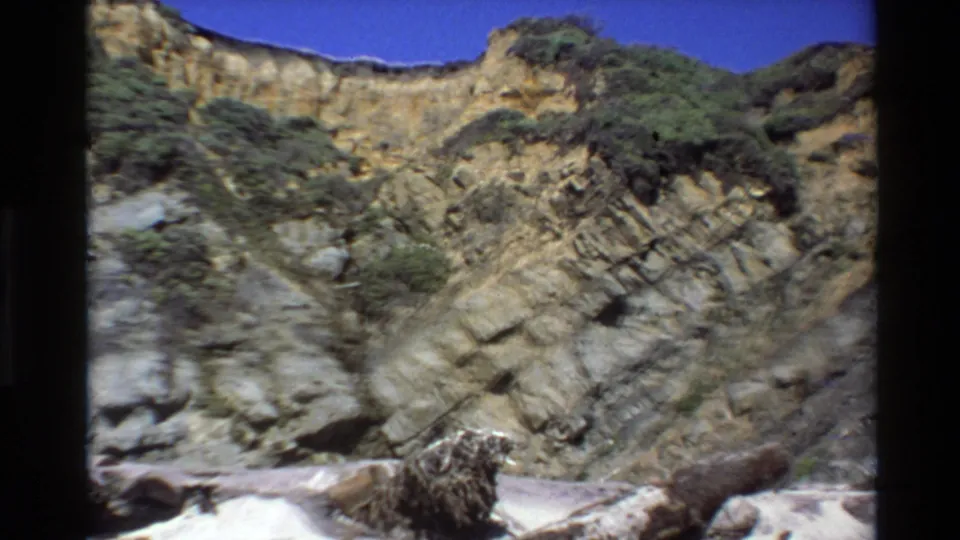 8mm Big Sur frame showing a closer section of layered cliff, scrubby plants, and pale beach or shoreline material below.