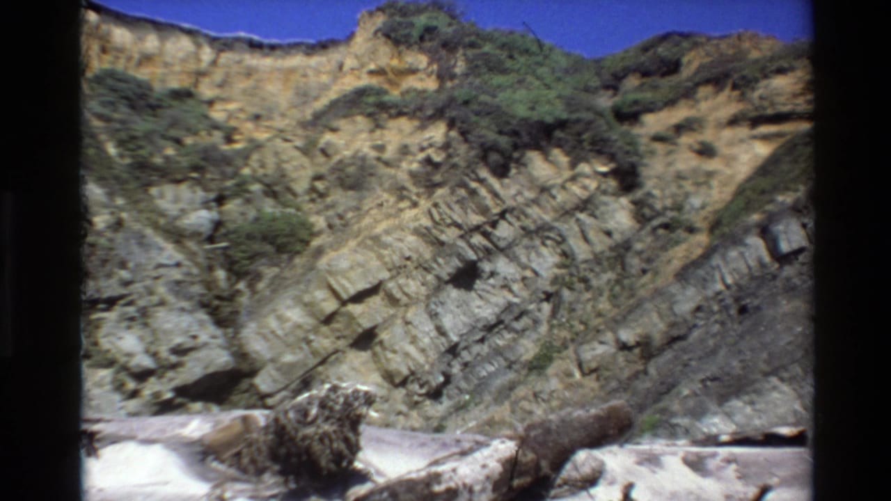 Final Big Sur frame with diagonal rock layers, green hillside shrubs, clear blue sky, and driftwood-like material in the foreground.