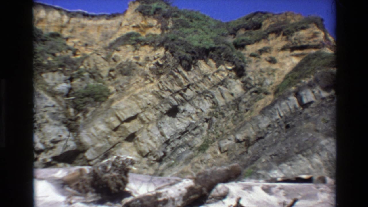 1980 8mm Big Sur coastline view with stratified cliffs, sparse vegetation, blue sky and driftwood on the beach.
