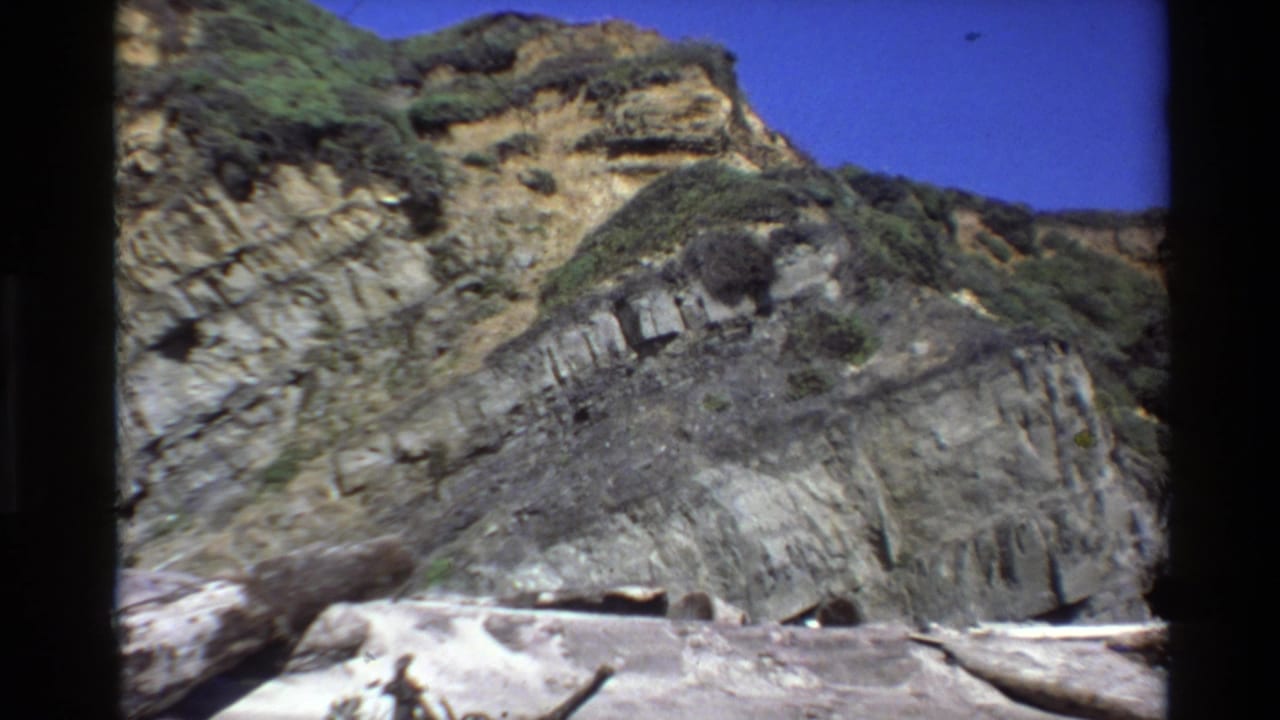 Vintage 8mm Big Sur coastal frame showing angled rock formations, sparse greenery, bright sky and driftwood on the beach.