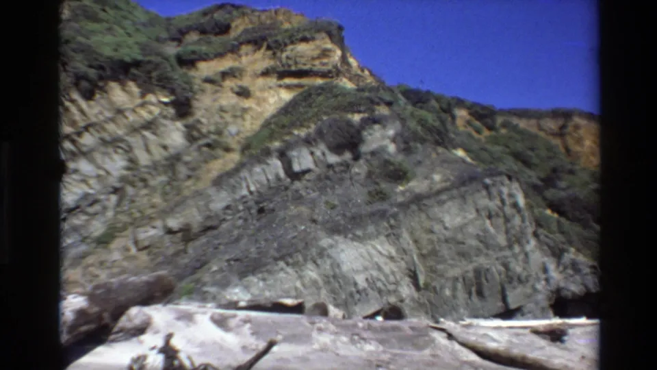 Big Sur coastal cliff in 1980 8mm footage, with layered rock, green vegetation, sandy beach, driftwood, and clear blue sky.