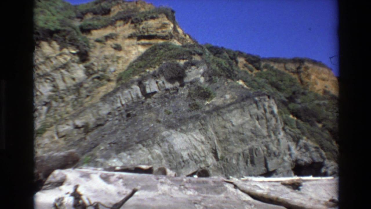 Big Sur cliff and beach view in 1980 8mm footage, showing weathered rock layers, low vegetation, driftwood, and bright sky.