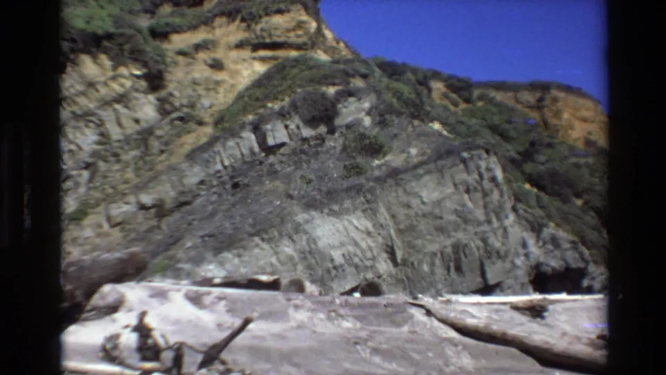 Closer Big Sur 8mm view of a sloping layered rock face, sparse vegetation, beach sand, and driftwood under blue sky.