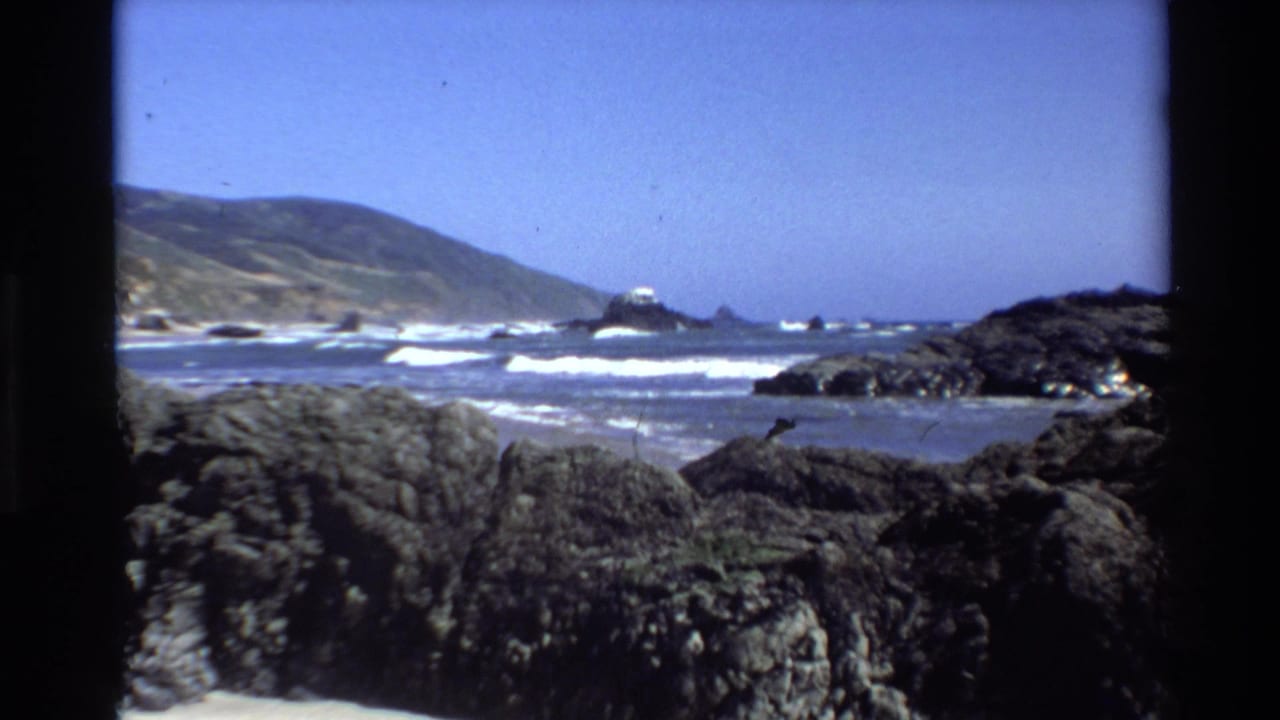 1980 8mm view of Big Sur, California, with dark rocks in front, white surf, coastal hills, and a small offshore island or sea stack.
