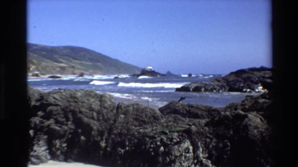 1980 8mm view of Big Sur, California, with dark rocks in front, white surf, coastal hills, and a small offshore island or sea stack.