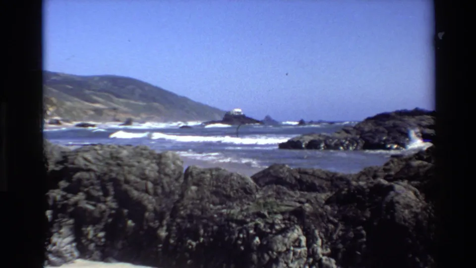 Big Sur coastal home-movie frame showing breaking waves, black foreground rocks, a distant sea stack, green-brown hills, and clear sky.