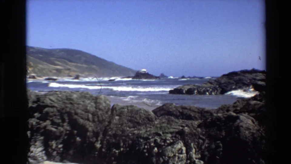 1980 Big Sur shoreline view with large foreground rocks, low surf bands, a small offshore formation, coastal hills, and open sky.