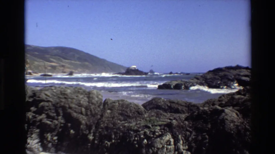 Big Sur, California, home-movie still with foreground rocks, Pacific surf, an offshore sea stack, sloping hills, and no visible people.