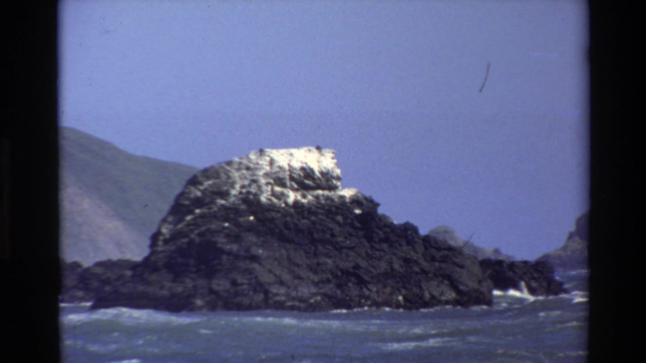 1980 Big Sur 8mm frame showing a dark rocky sea stack, blue water, white surf, and a distant coastal slope under clear sky.