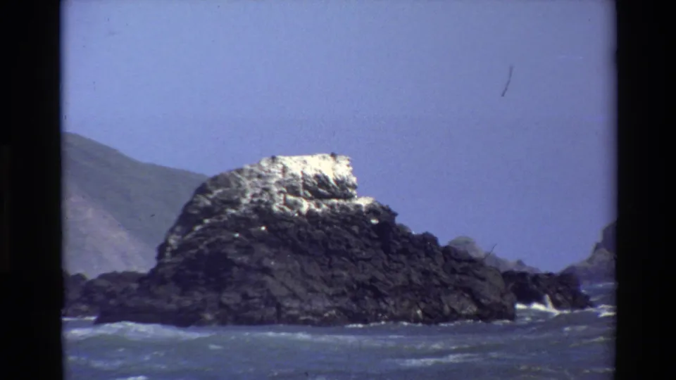 1980 Big Sur 8mm frame showing a dark rocky sea stack, blue water, white surf, and a distant coastal slope under clear sky.