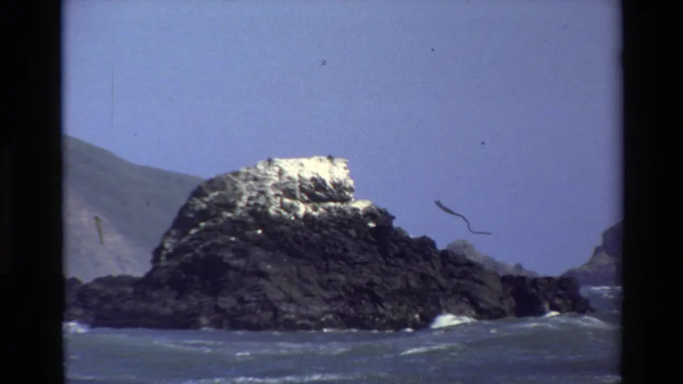 Big Sur coastal frame from 1980 showing a dark offshore rock with a pale top, surrounding surf, blue sky, and visible film artifacts.