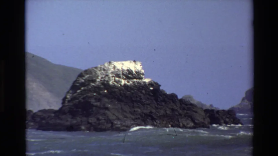 1980 Big Sur home-movie frame holding on a dark sea stack, white wave lines, blue water, and a muted coastal hillside.