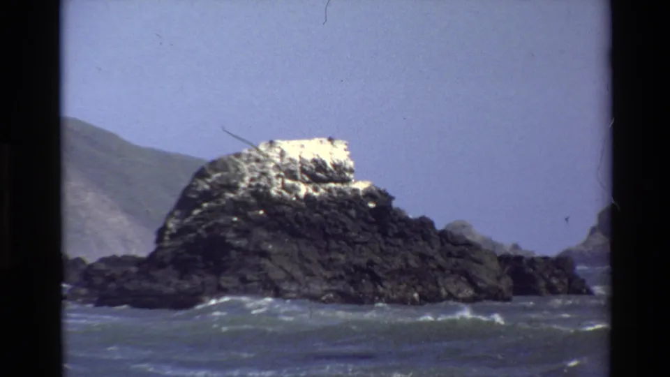 Big Sur 1980 8mm frame showing the offshore rock formation, moving surf, open blue sky, and distant shoreline at the frame edges.