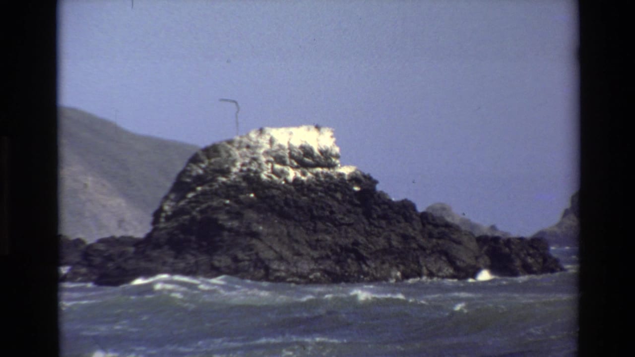 1980 Big Sur home-movie frame showing a dark coastal rock rising from choppy blue-gray water with hazy hills behind it.