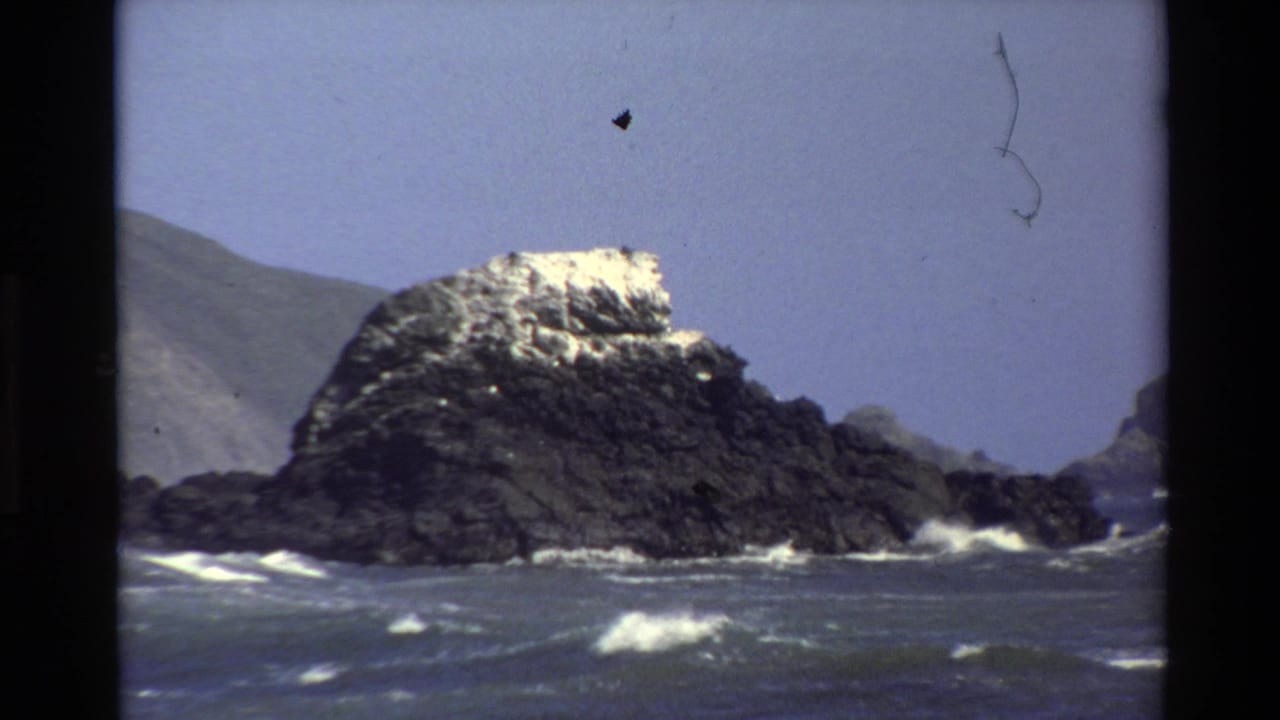 Large dark sea rock with waves breaking at its base and hills in the background.
