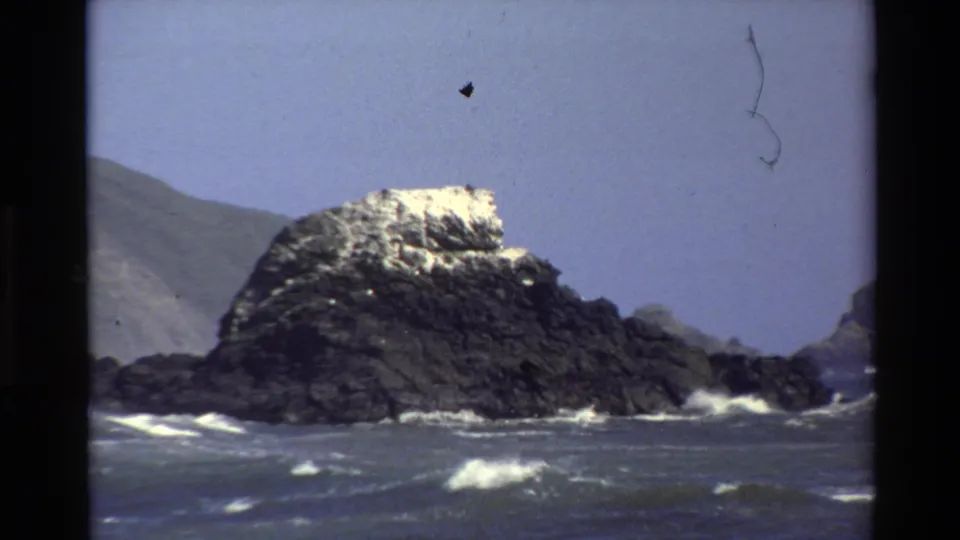 Big Sur coastal frame from 1980 showing waves striking a dark offshore rock beneath a muted blue sky.