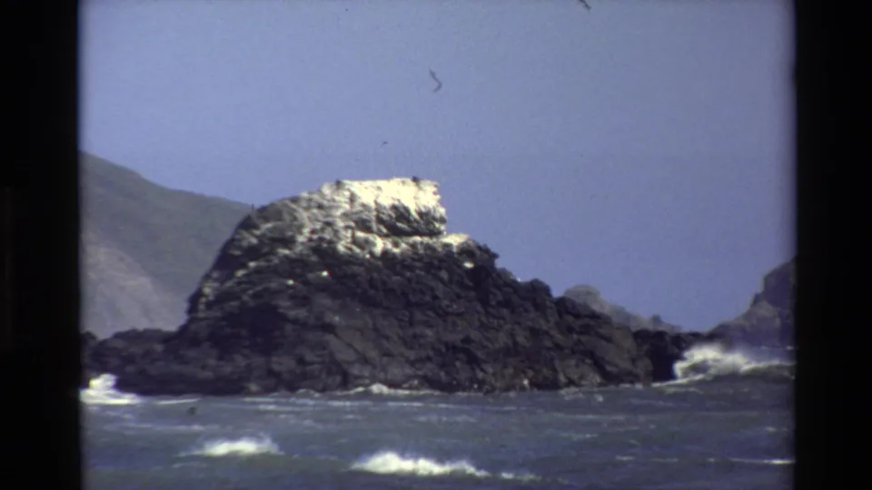 Vintage 8mm view of a Big Sur offshore rock with white surf moving across the foreground water.