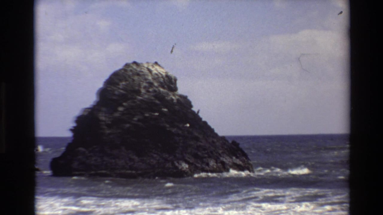1980 Big Sur 8mm frame showing a large offshore rock, blue ocean, white surf, and soft partly cloudy sky.
