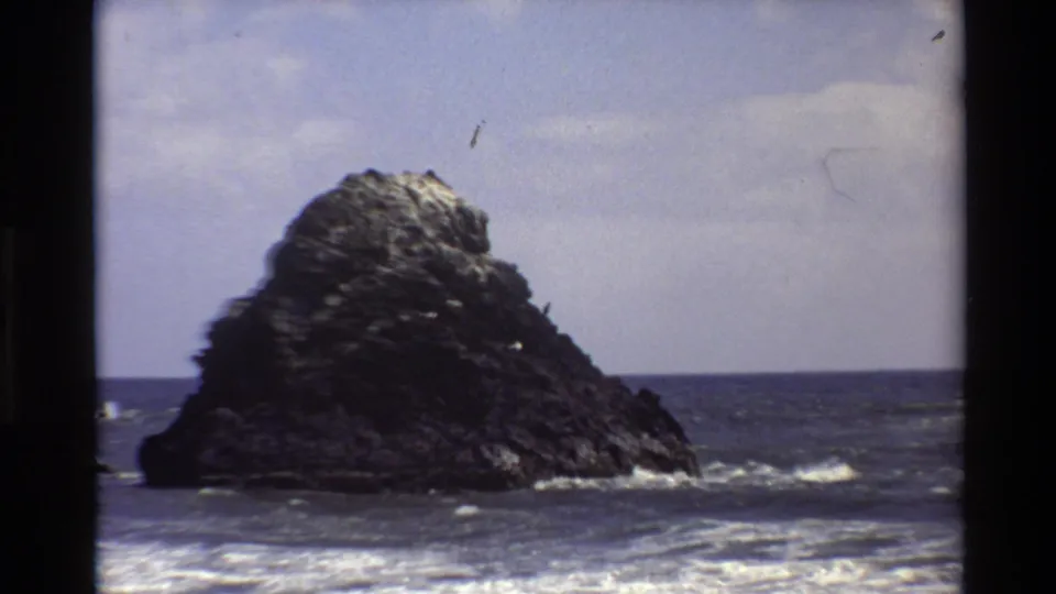 1980 Big Sur 8mm frame showing a large offshore rock, blue ocean, white surf, and soft partly cloudy sky.