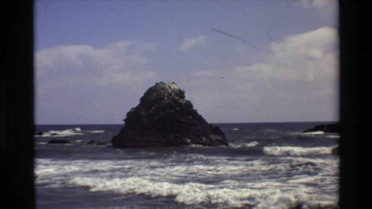Offshore rock centered beyond rolling waves with smaller rocks near the horizon.