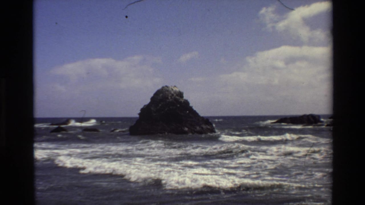 1980 Big Sur home-movie frame with the offshore rock set farther back behind wide bands of surf and ocean foam.