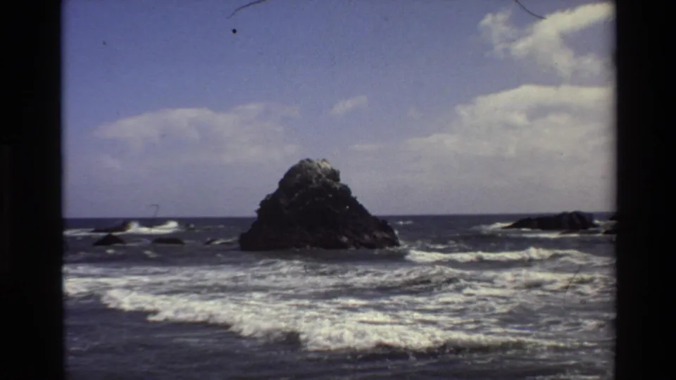 1980 Big Sur home-movie frame with the offshore rock set farther back behind wide bands of surf and ocean foam.