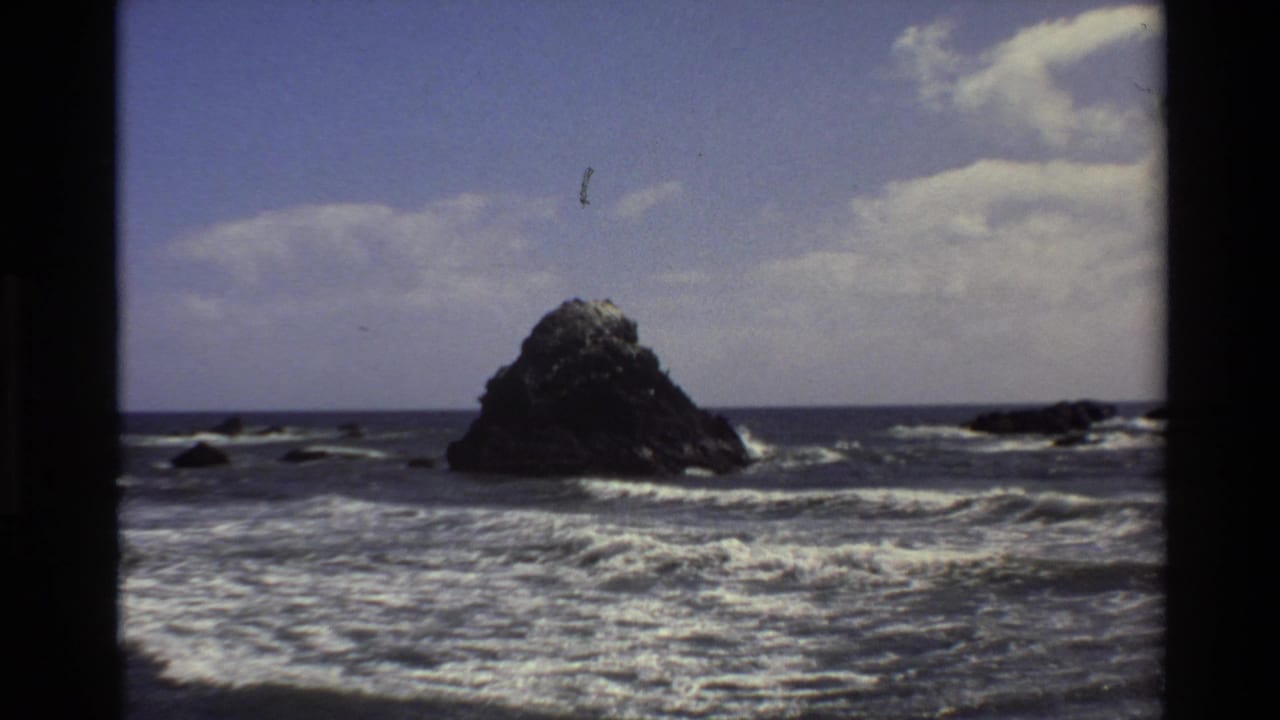 1980 Big Sur 8mm frame showing surf breaking around a large dark offshore rock under blue sky and clouds.