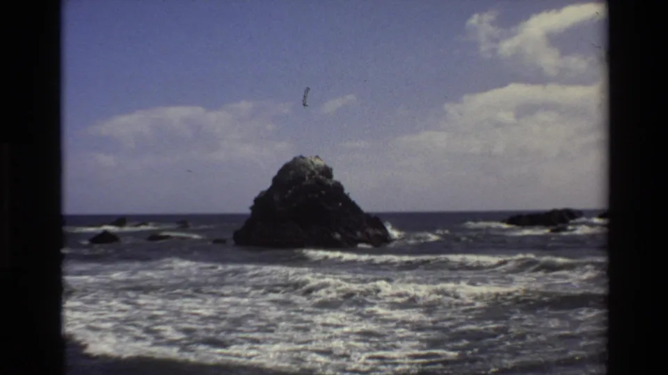 1980 Big Sur 8mm frame showing surf breaking around a large dark offshore rock under blue sky and clouds.