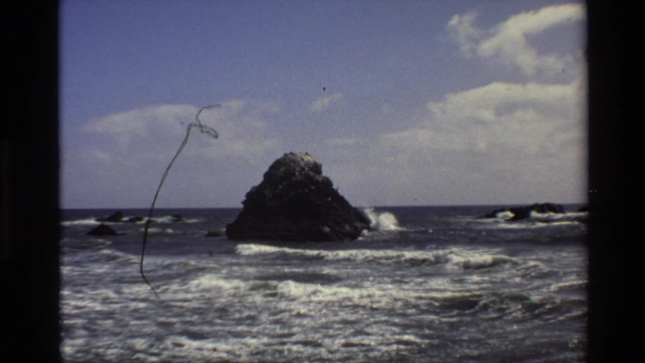 Large rock offshore with waves breaking nearby and film scratches over the sky.