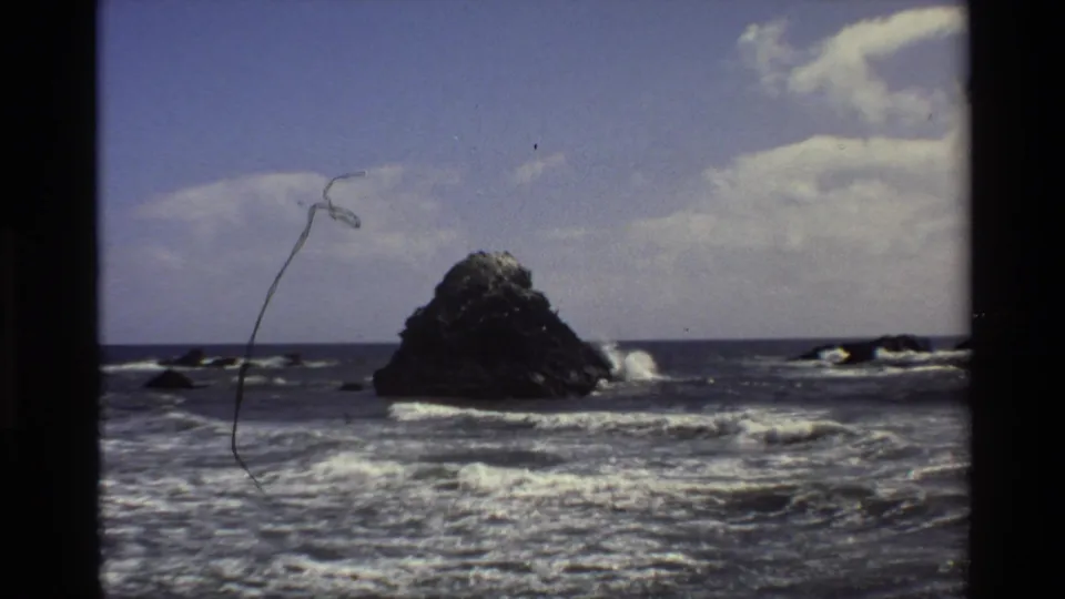 Big Sur coastal frame with a dark sea stack, breaking surf, clouded sky, and visible 8mm film marks.