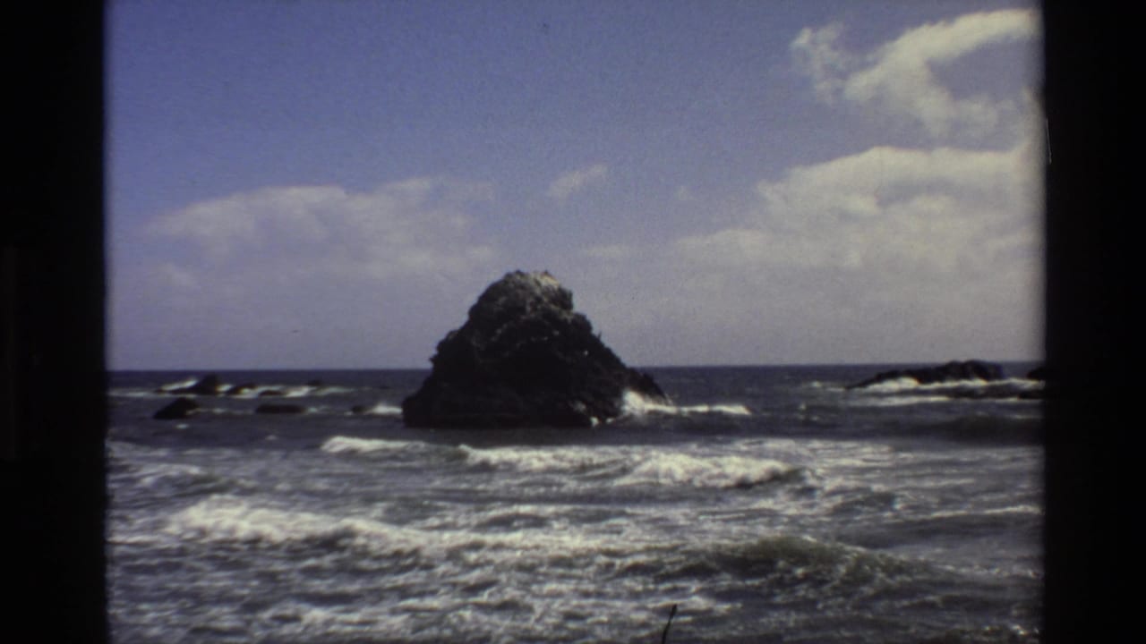 1980 Big Sur home-movie frame showing a rocky offshore formation beyond choppy water and low foamy breakers.