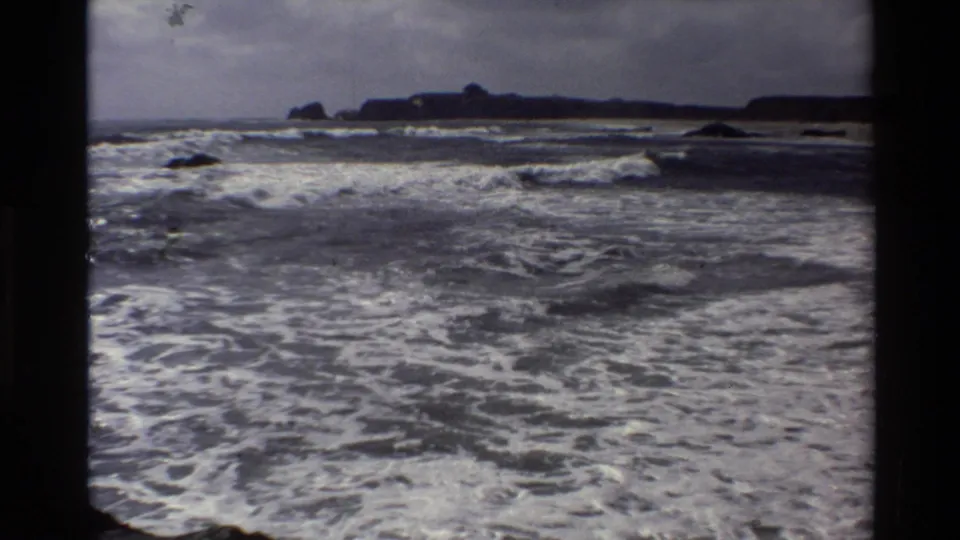 Big Sur 8mm frame with foamy shoreline water, distant rocks, and a low coastal horizon under a cloudy sky.