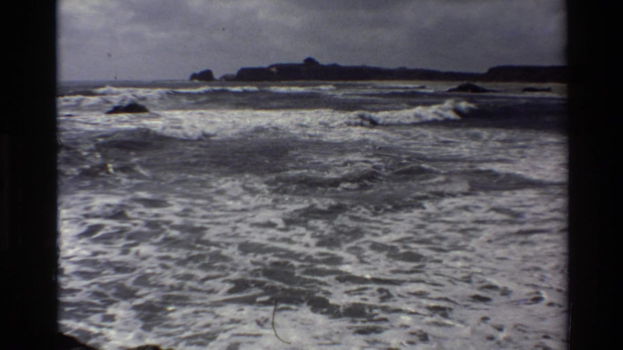 1980 8mm Big Sur coastal footage shows turbulent Pacific surf, white foam, dark rocks, and a distant headland within black film borders.