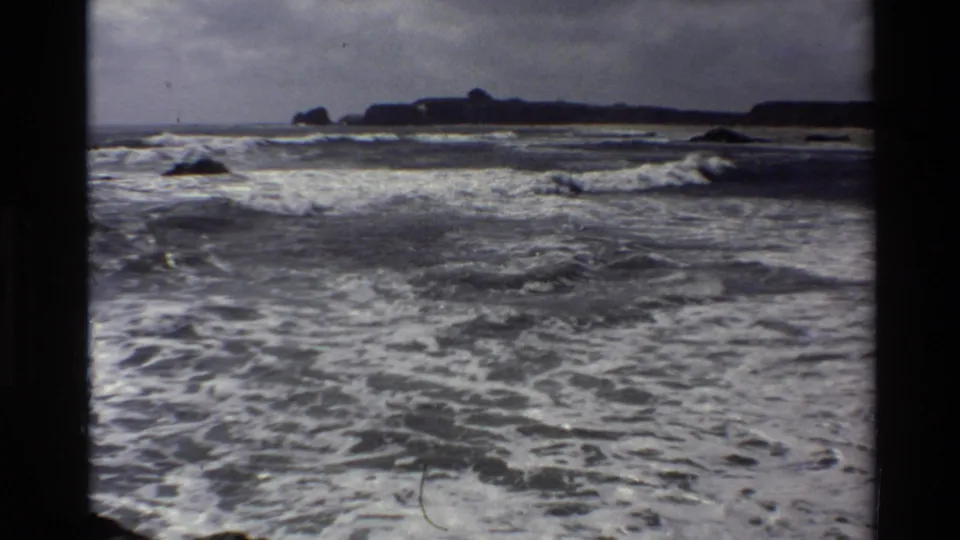 1980 8mm Big Sur coastal footage shows turbulent Pacific surf, white foam, dark rocks, and a distant headland within black film borders.