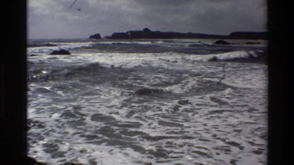 The Big Sur shoreline appears through an 8mm frame as white-capped waves churn below a gray sky and dark rocky coastline.