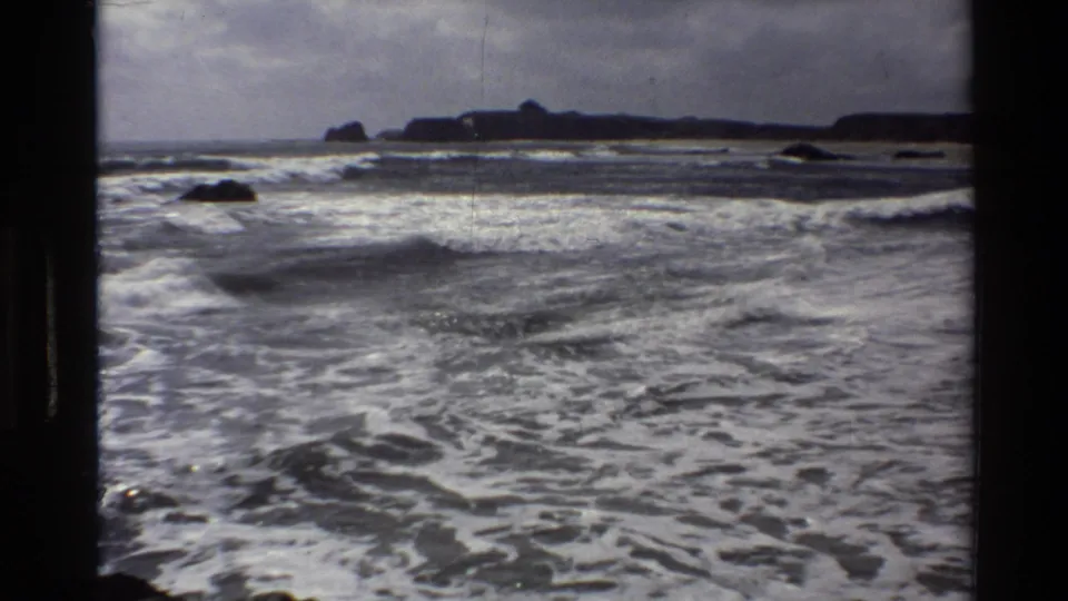 Vintage 8mm footage from Big Sur shows lower rolling surf, scattered rocks, and a dark headland across the horizon.
