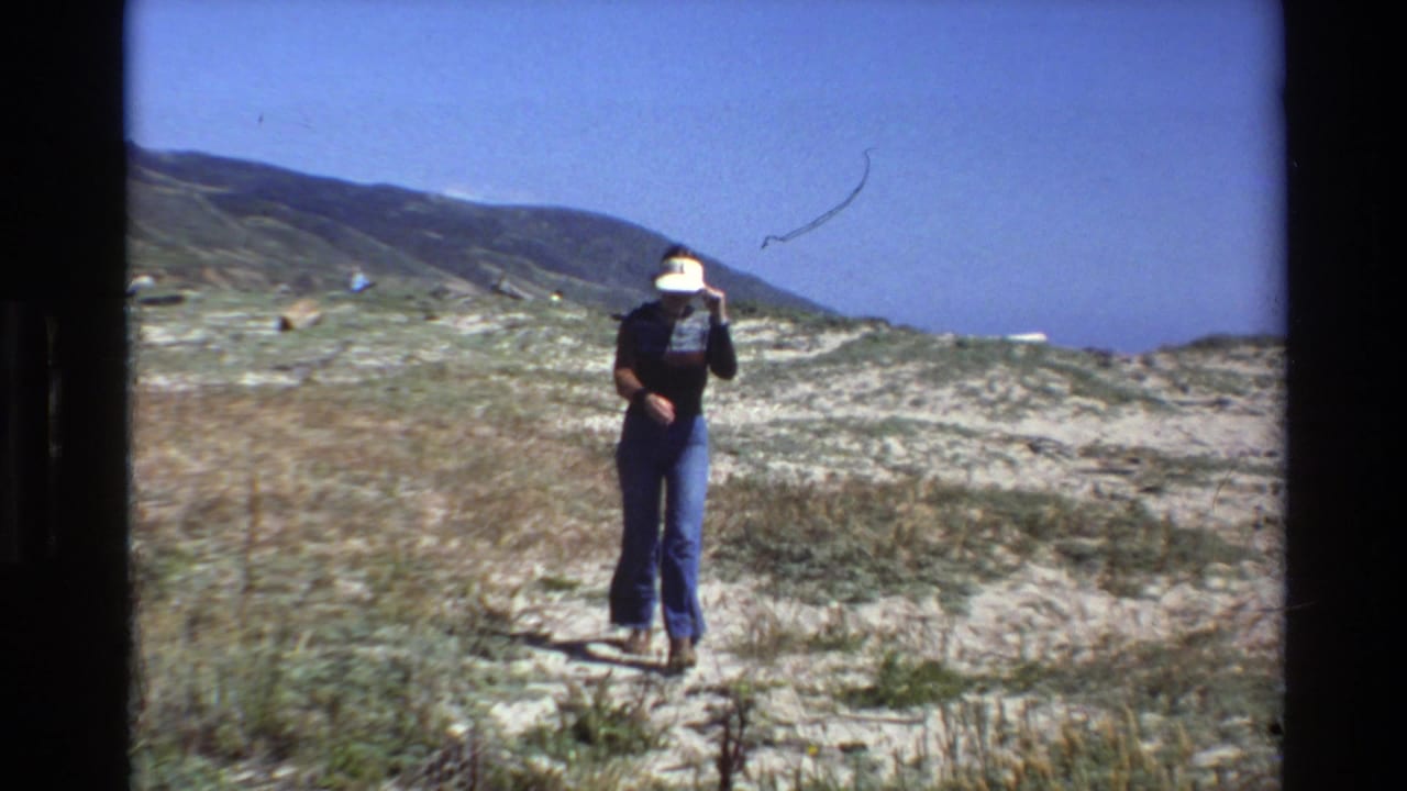 1980 Big Sur home-movie frame showing a distant person in a white visor, dark top, and blue jeans walking through sandy coastal dunes.