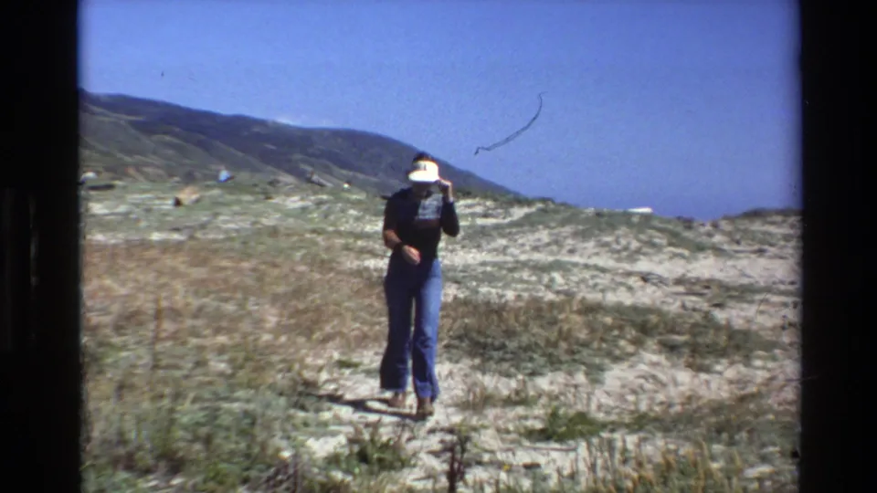 1980 Big Sur home-movie frame showing a distant person in a white visor, dark top, and blue jeans walking through sandy coastal dunes.