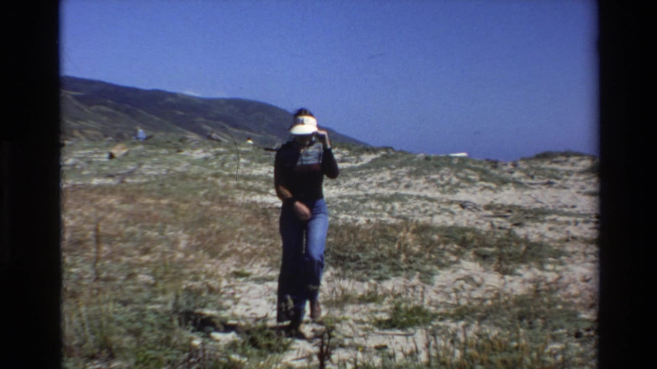 Person walking in dune grass with hills and clear sky behind them.