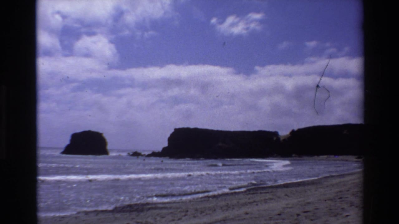1980 Big Sur 8mm frame showing surf, beach, dark coastal cliffs, and a sea stack under partly cloudy sky.