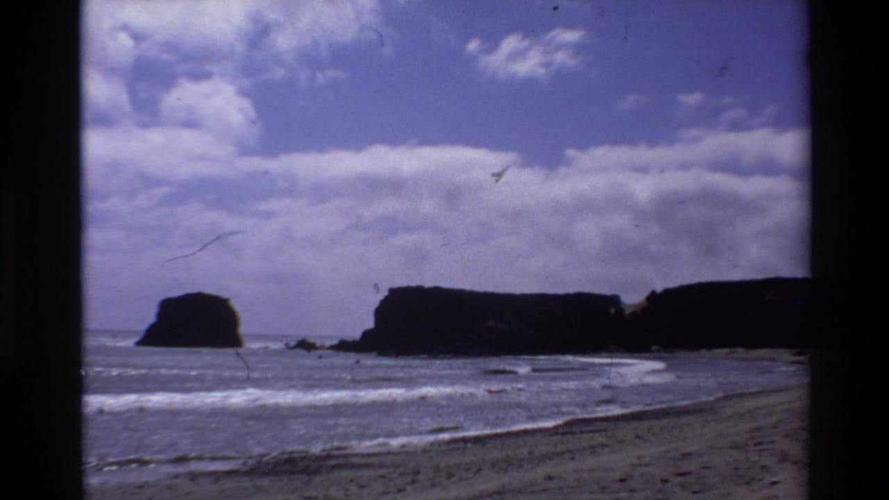 1980 Big Sur home-movie frame of waves, beach, dark headlands, offshore rocks, and partly cloudy sky.