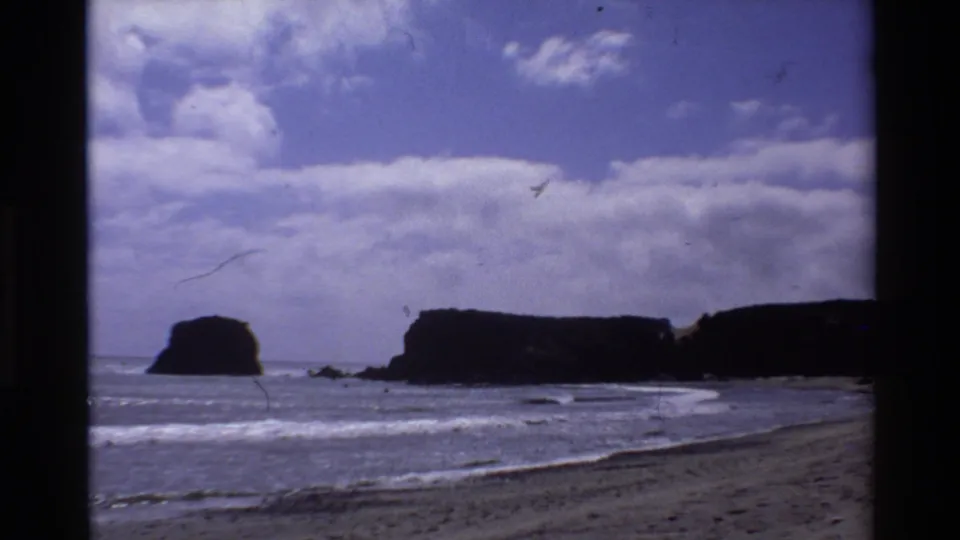 1980 Big Sur home-movie frame of waves, beach, dark headlands, offshore rocks, and partly cloudy sky.