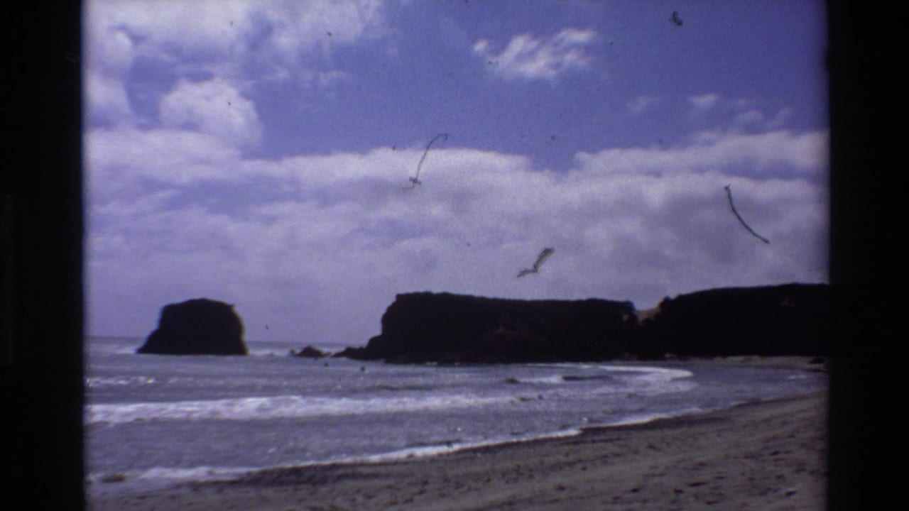 8mm home movie still, c. 1980, Big Sur California — beach foreground, silhouetted rock formations offshore, partly cloudy sky, film scratches visible.