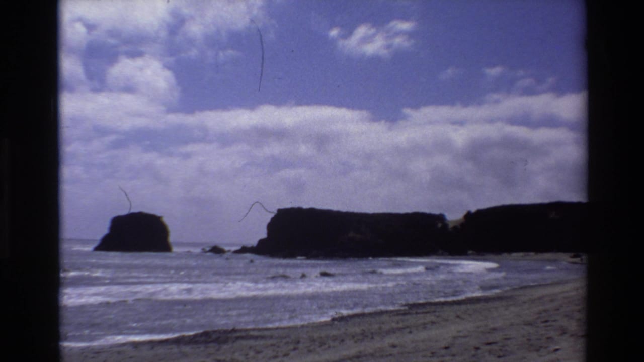 Coastal beach scene with dark sea stacks, rolling waves, and blue cloudy sky; 8mm with film hair artifact.