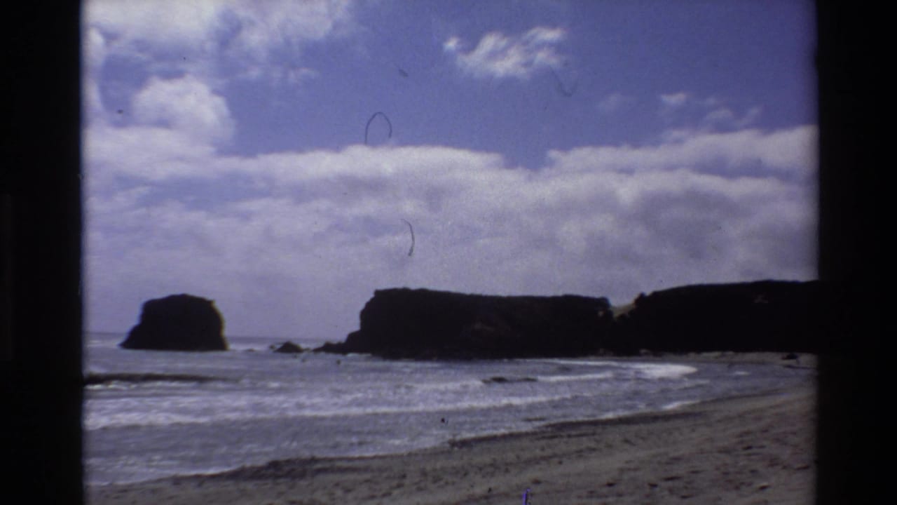 8mm home movie still, c. 1980, Big Sur California — wider framing with more sky, sea stacks lower in frame, wet sand foreground, film scratches visible.