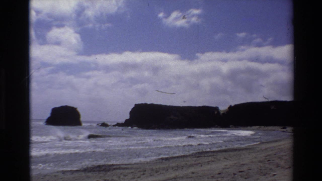 8mm home movie still, c. 1980, Big Sur California — sea stacks silhouetted at left-center, sandy beach foreground, partly cloudy sky occupying upper half of frame.