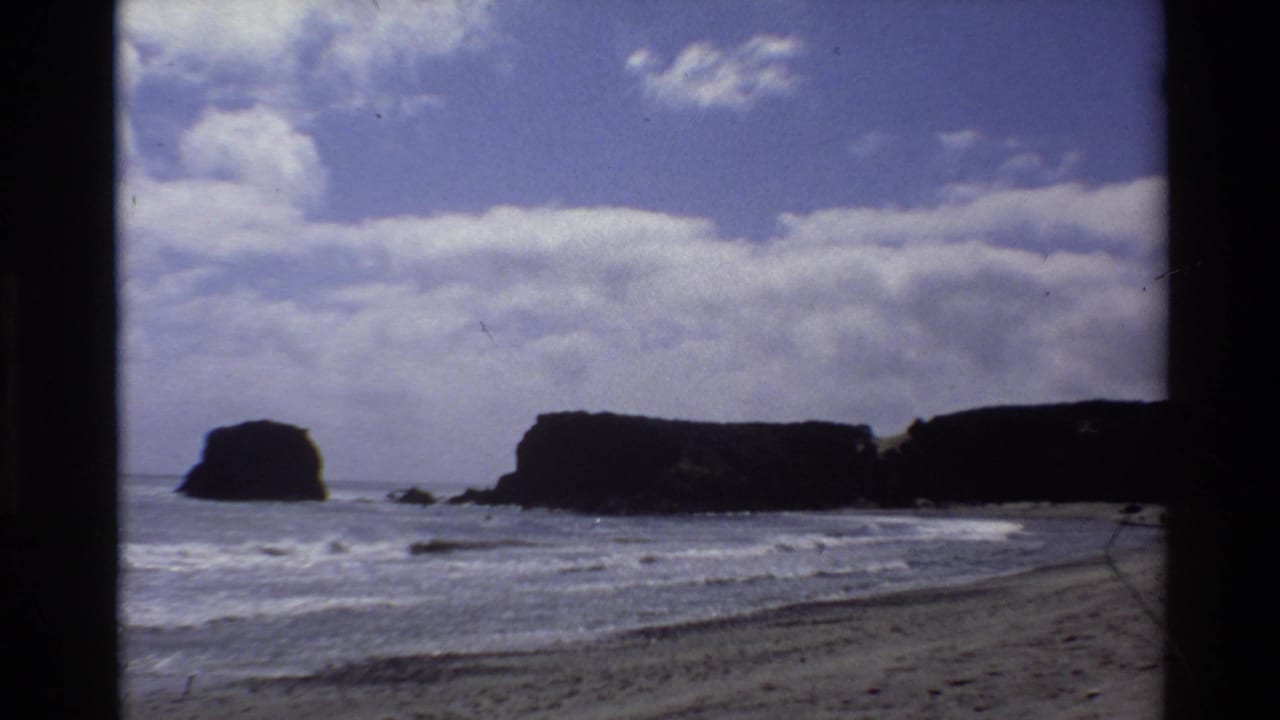 Super 8 wide shot of a sandy California coastal beach circa 1980, with two prominent dark sea stacks and cumulus clouds.