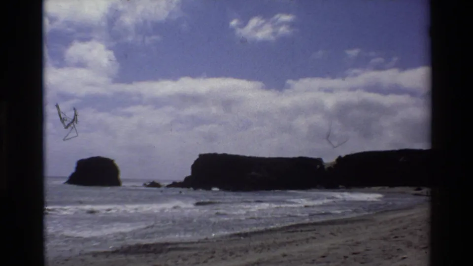 Super 8 frame of Big Sur coast circa 1980, showing sea stacks and beach with film scratch artifacts across the upper portion.