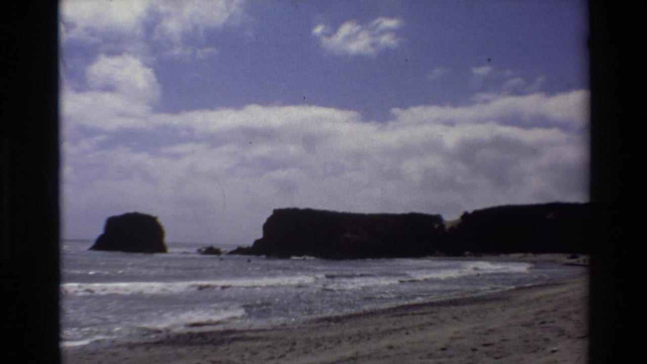 Super 8 coastal frame from Big Sur circa 1980, showing one prominent sea stack, sandy beach, and cumulus cloud sky.