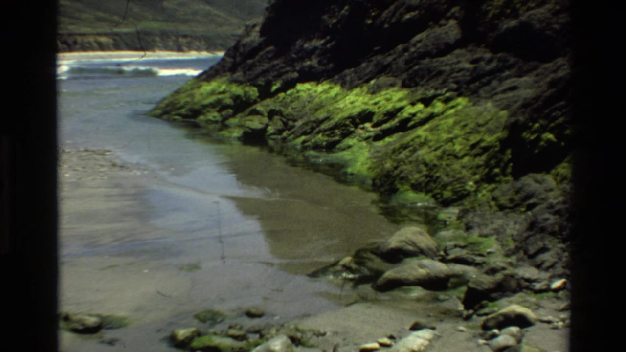Super 8 close coastal shot likely from Big Sur circa 1980, showing a moss-covered rocky cliff, narrow tidal cove, and rounded boulders.
